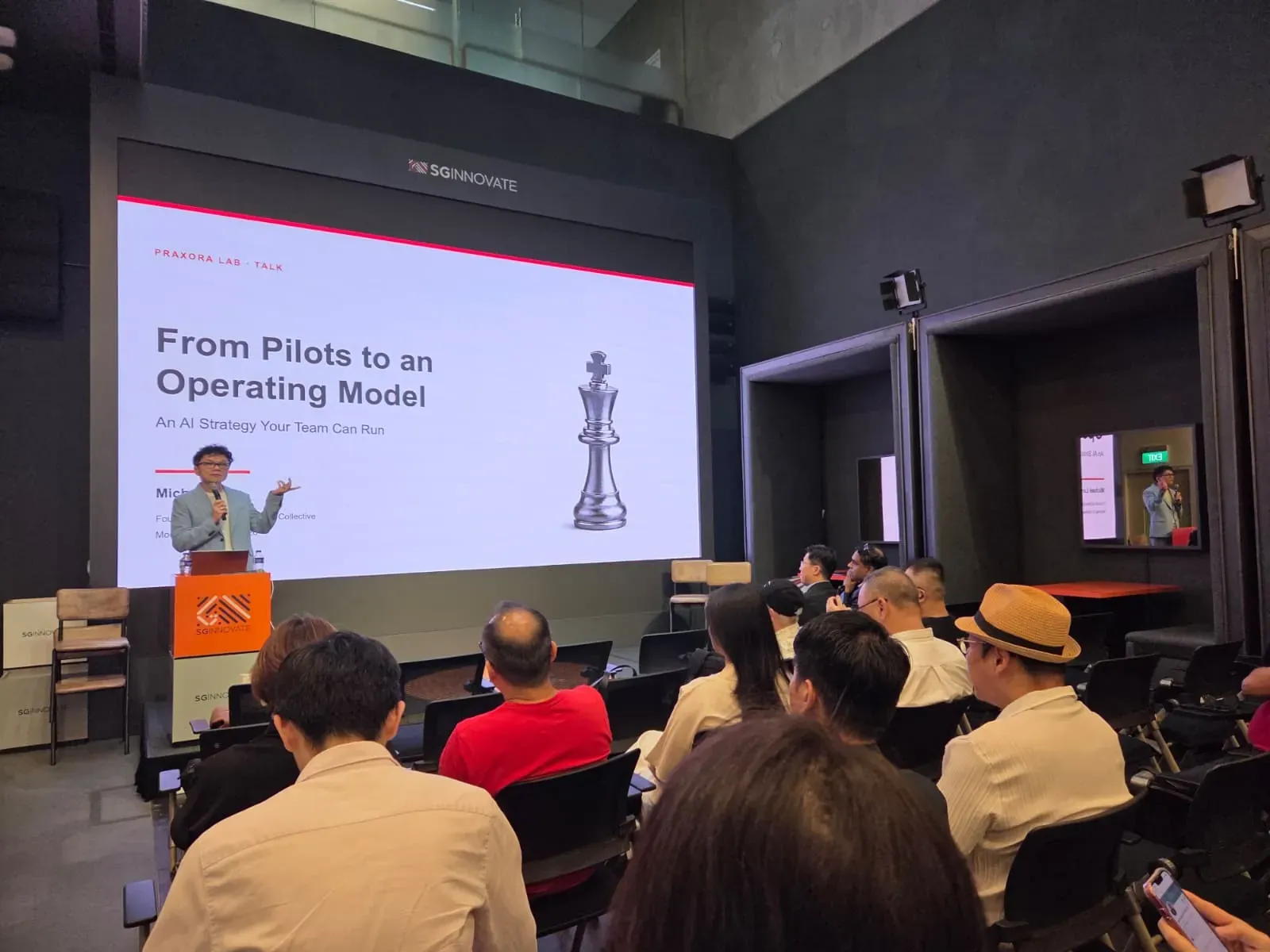 Michael Low at the lectern beside a screen reading From Pilots to an Operating Model, seen over the heads of the seated audience.